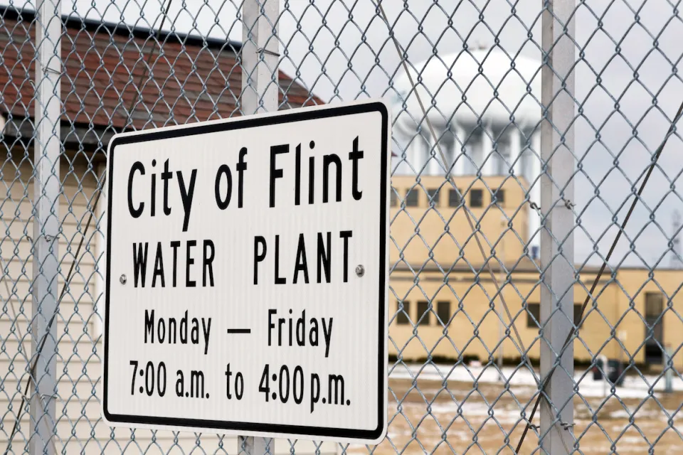 Close-up of sign on chain link fence "City of Flint Water Plant" with hours.